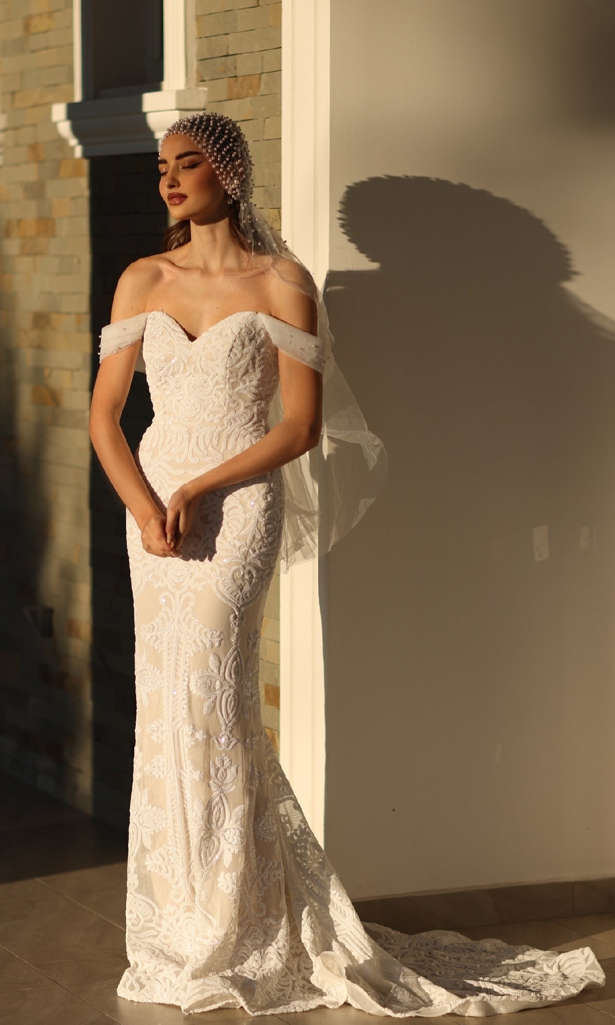 Wearing the Off-Shoulder White Bridal Gown: Tina Holly TW103 and a pearl headpiece, a woman stands in sunlight by a wall, her graceful shadow cast behind her.